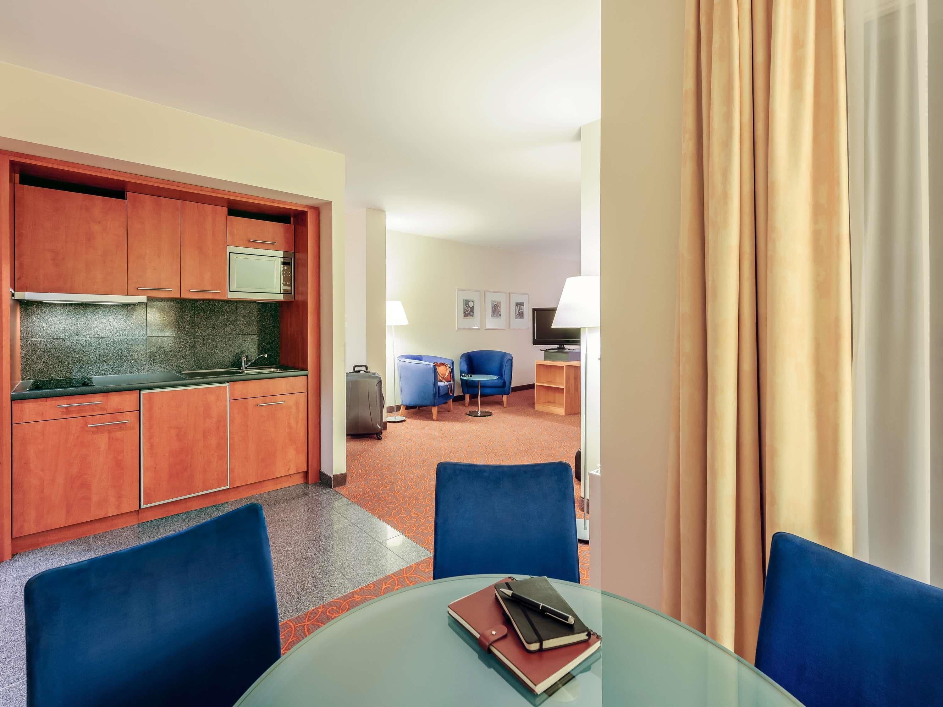 A hotel room with a kitchenette, a dining area with a glass table, and a living area with a TV and seating.