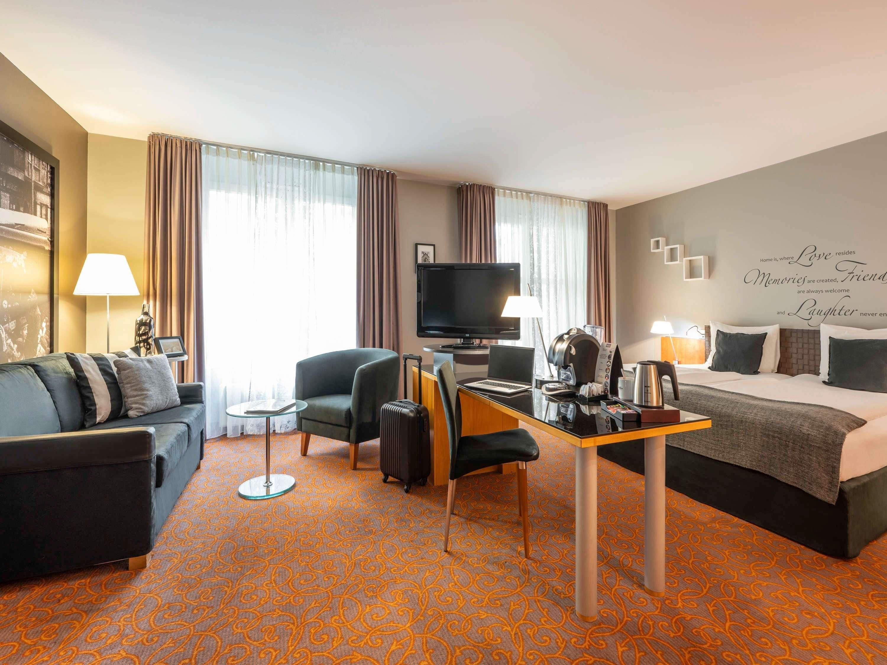 A hotel room with a sofa, armchair, desk with laptop, coffee maker, and a bed.
