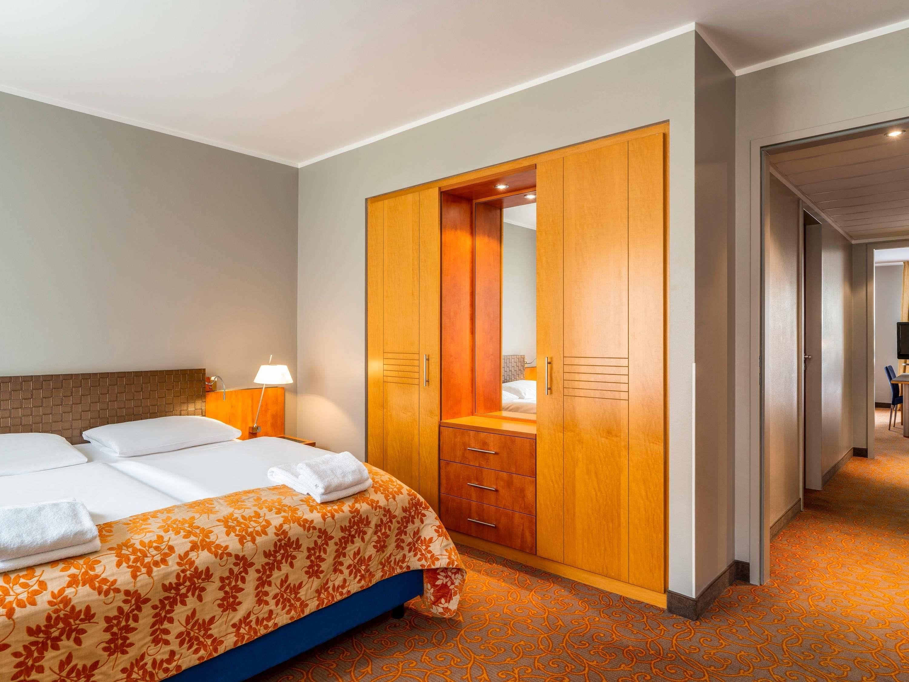 A hotel room with a bed, a wooden wardrobe, a desk, and a chair.
