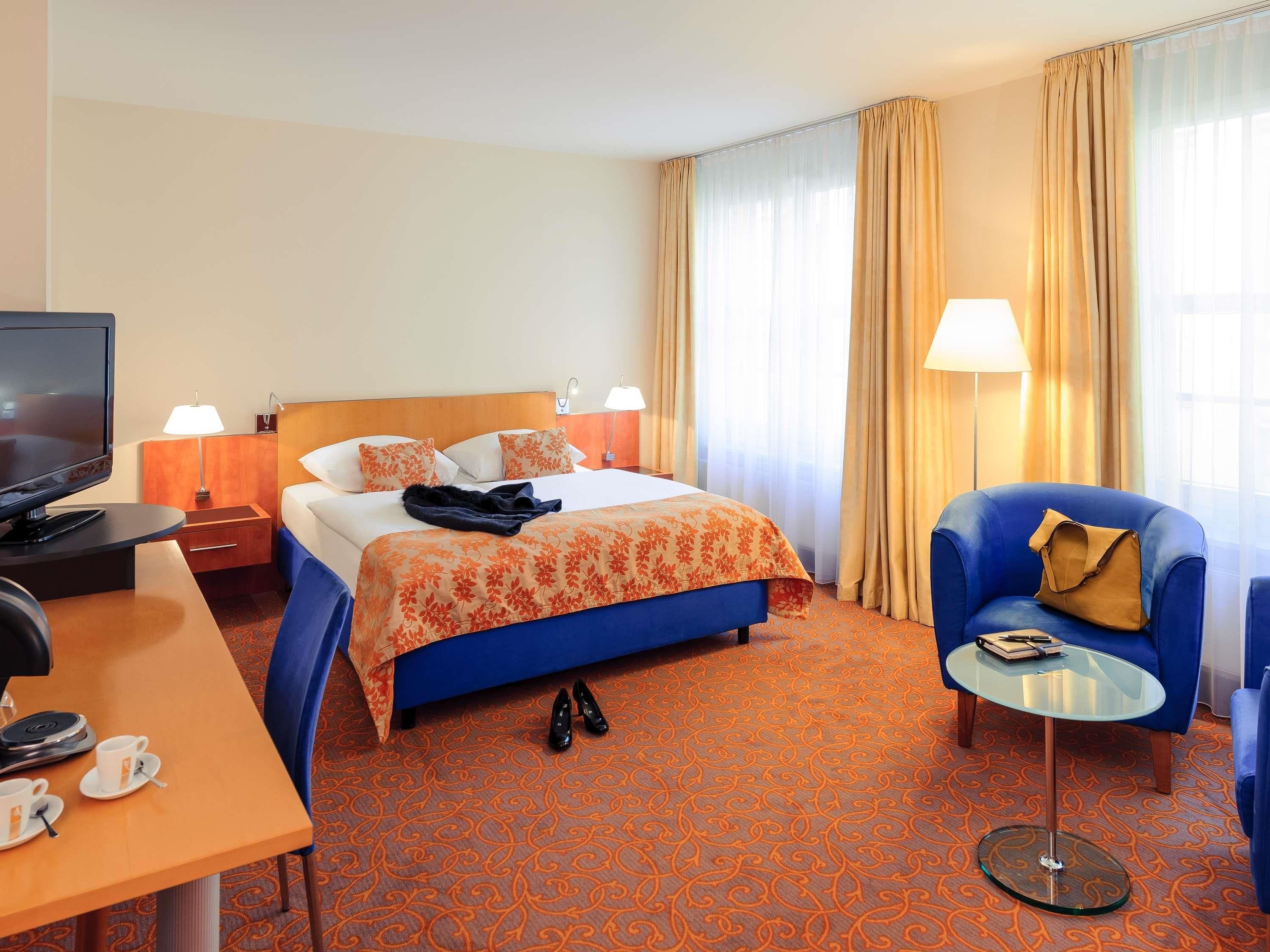 Habitación de hotel con cama, televisor, escritorio con silla, una mesita y un sillón azul.