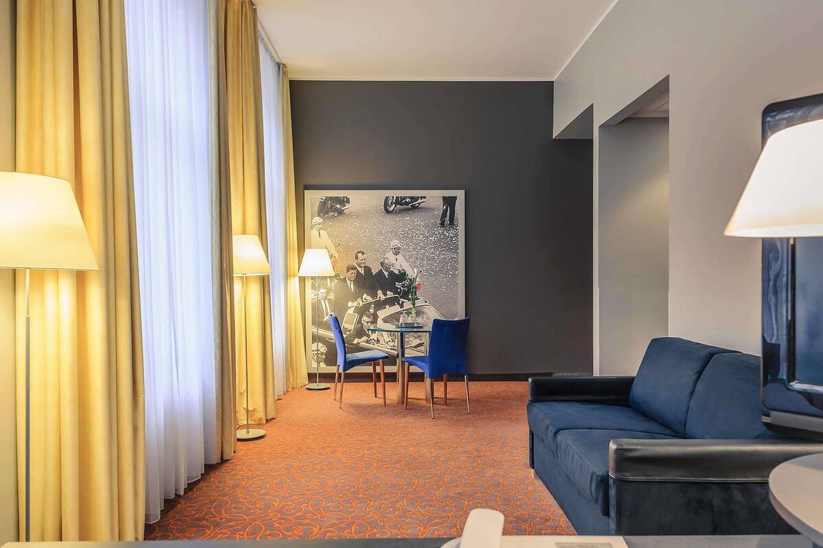 Hotellihuone, jossa on sininen sohva, lamppuvarustettu työpöytä, tuoli, suuri abstrakti maalaus ja kuviollinen matto.