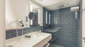 A modern bathroom with a white vanity, a large mirror, a glass shower enclosure, and dark blue tiled walls. - Mercure Hotel & Residenz Checkpoint Charlie (Berlin)