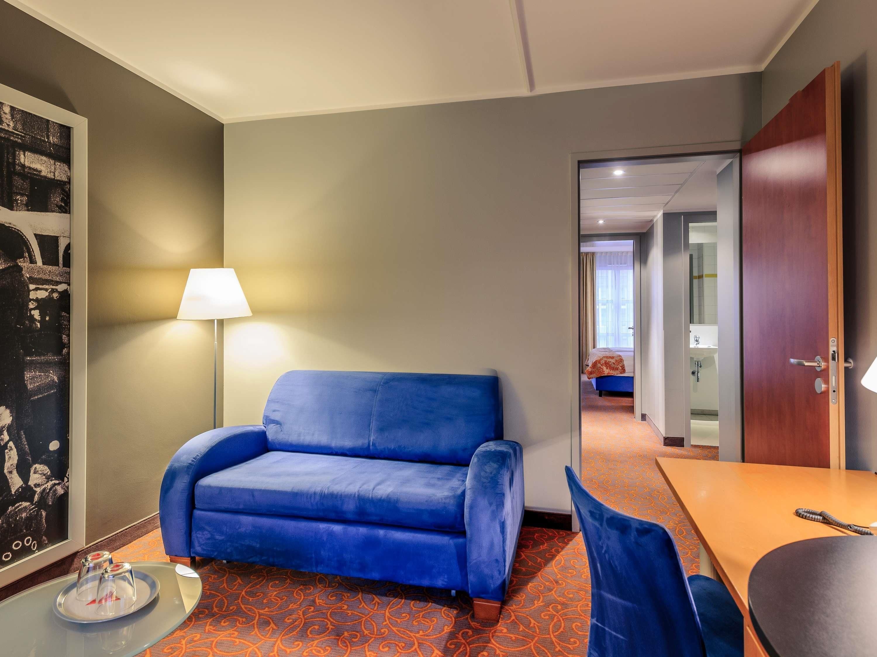 A hotel room with a blue sofa, a small table with a tray of glasses, a lamp, and a view into the bathroom.