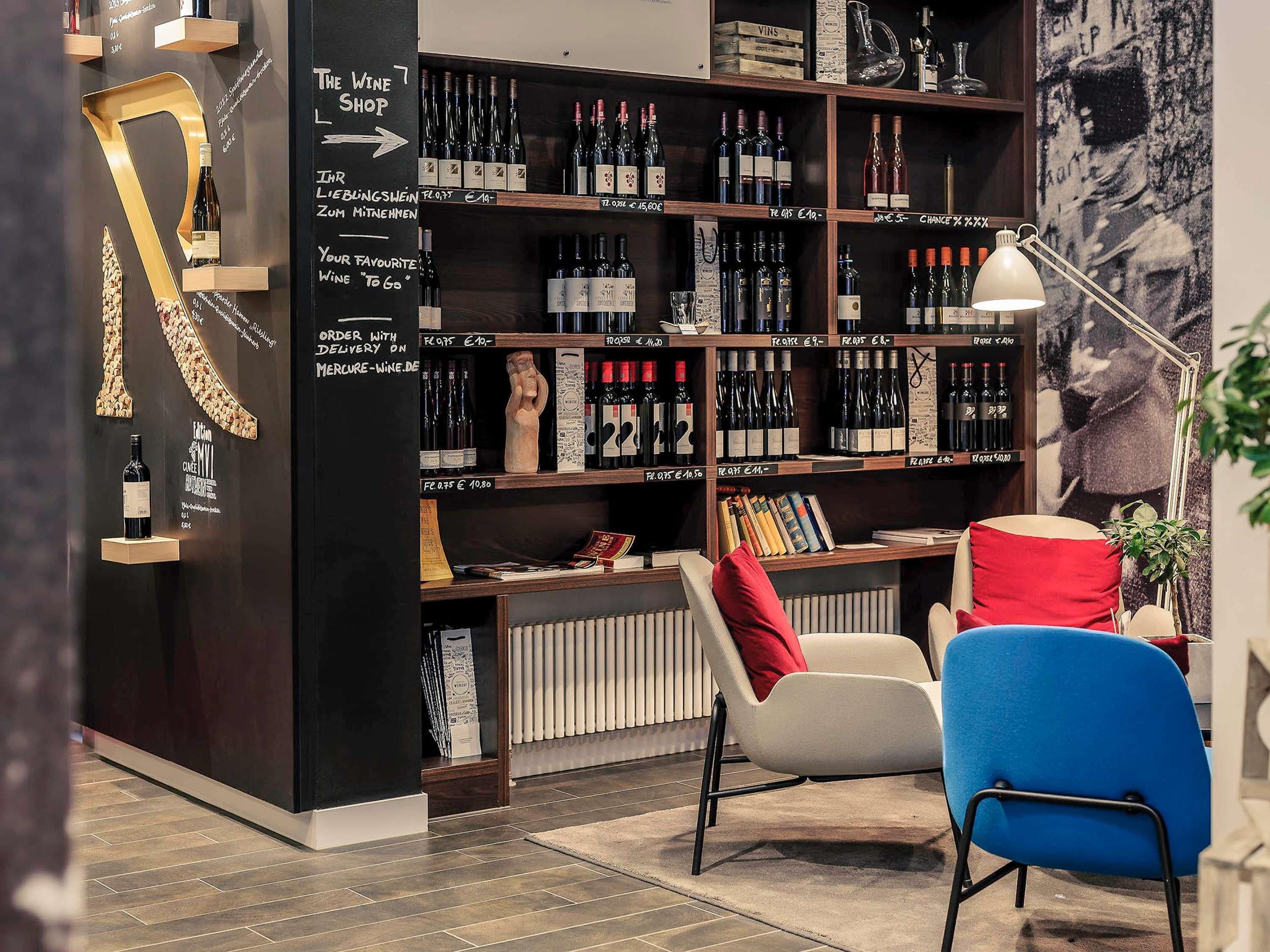 Interior de loja de vinhos com prateleiras cheias de garrafas, uma cadeira, uma mesa e uma parede com uma grande letra 'R'.