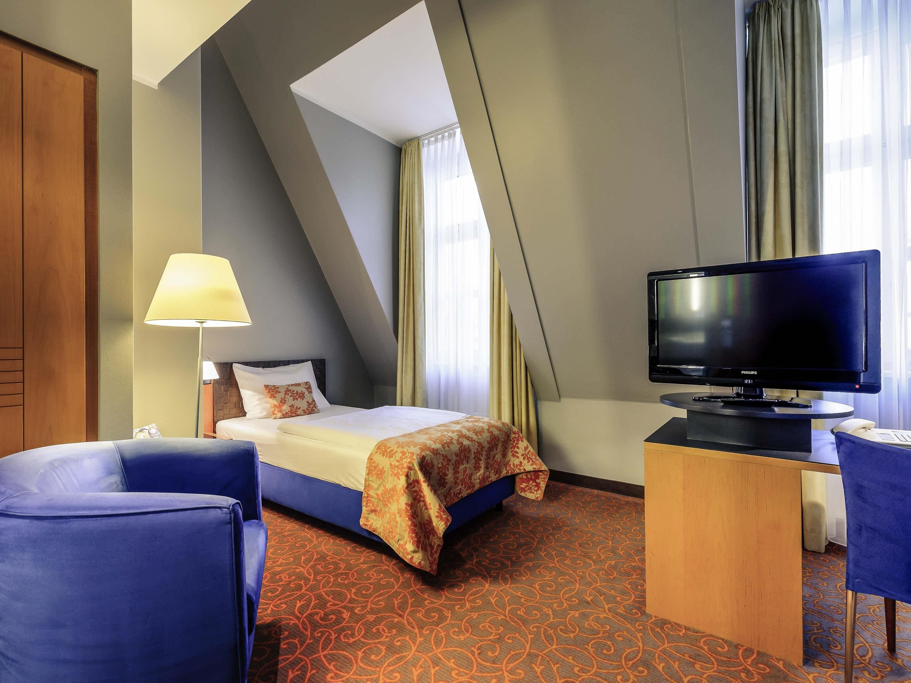 A hotel room with a bed, a desk with a television, a chair, and a lamp.