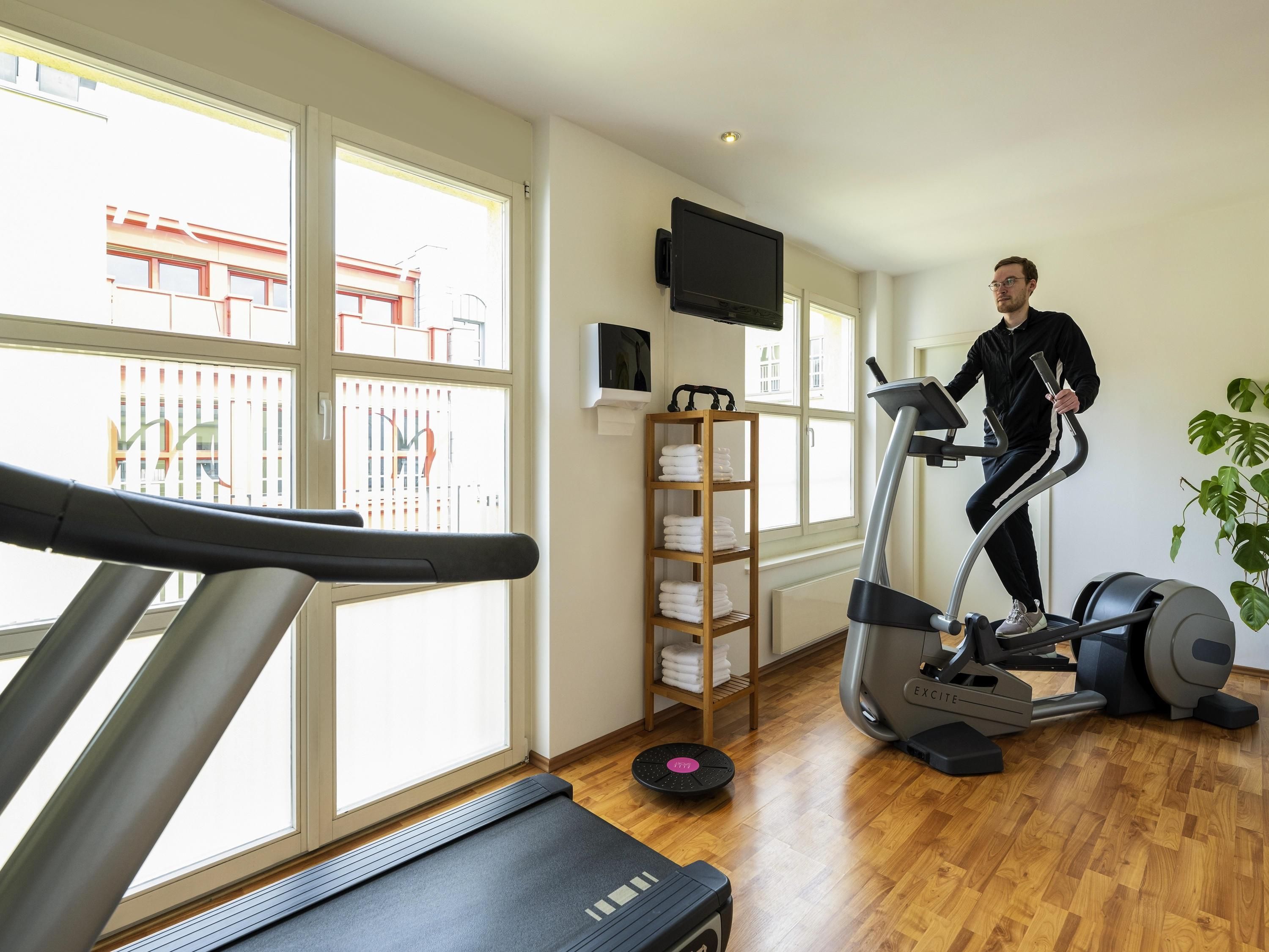 A gym with a treadmill, elliptical, and weights, large windows, and a TV on the wall.