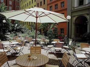 Un patio con mobiliario exterior, una sombrilla grande y un edificio con múltiples ventanas al fondo.