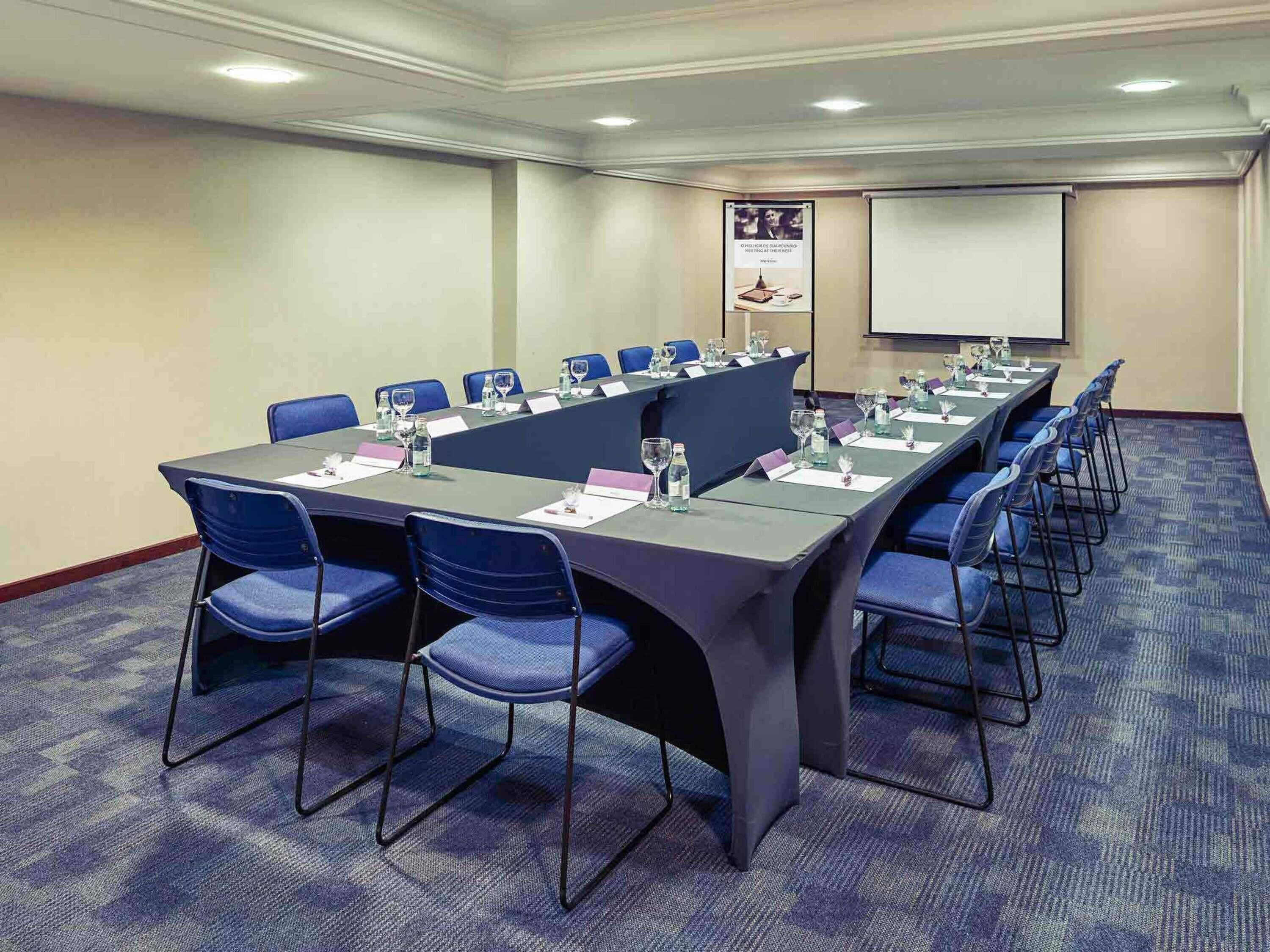 A conference room with a long table, blue chairs, name cards, water bottles, and a projector screen.