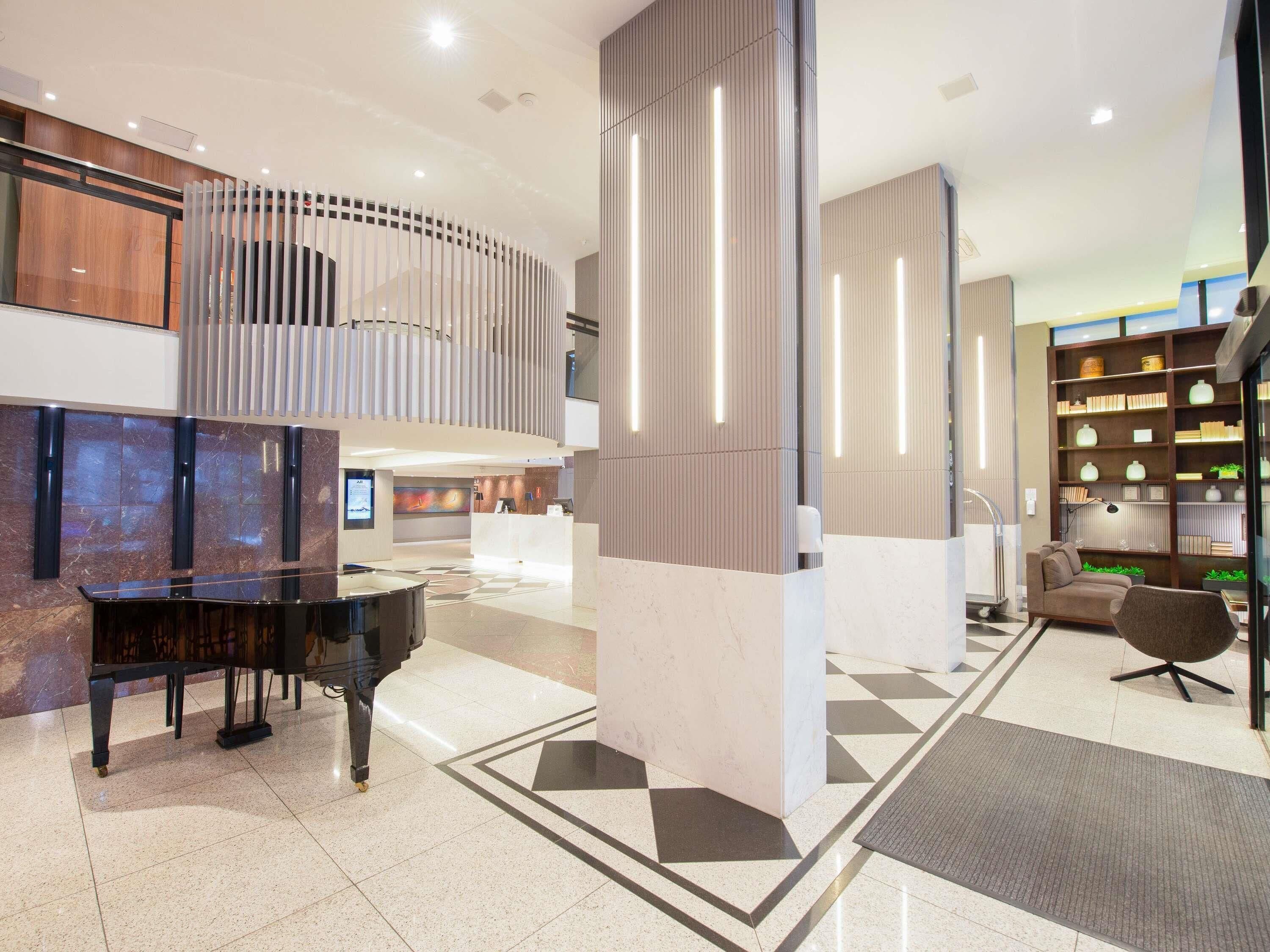 A modern lobby with a grand piano, a reception desk, a high ceiling, and a large bookshelf.