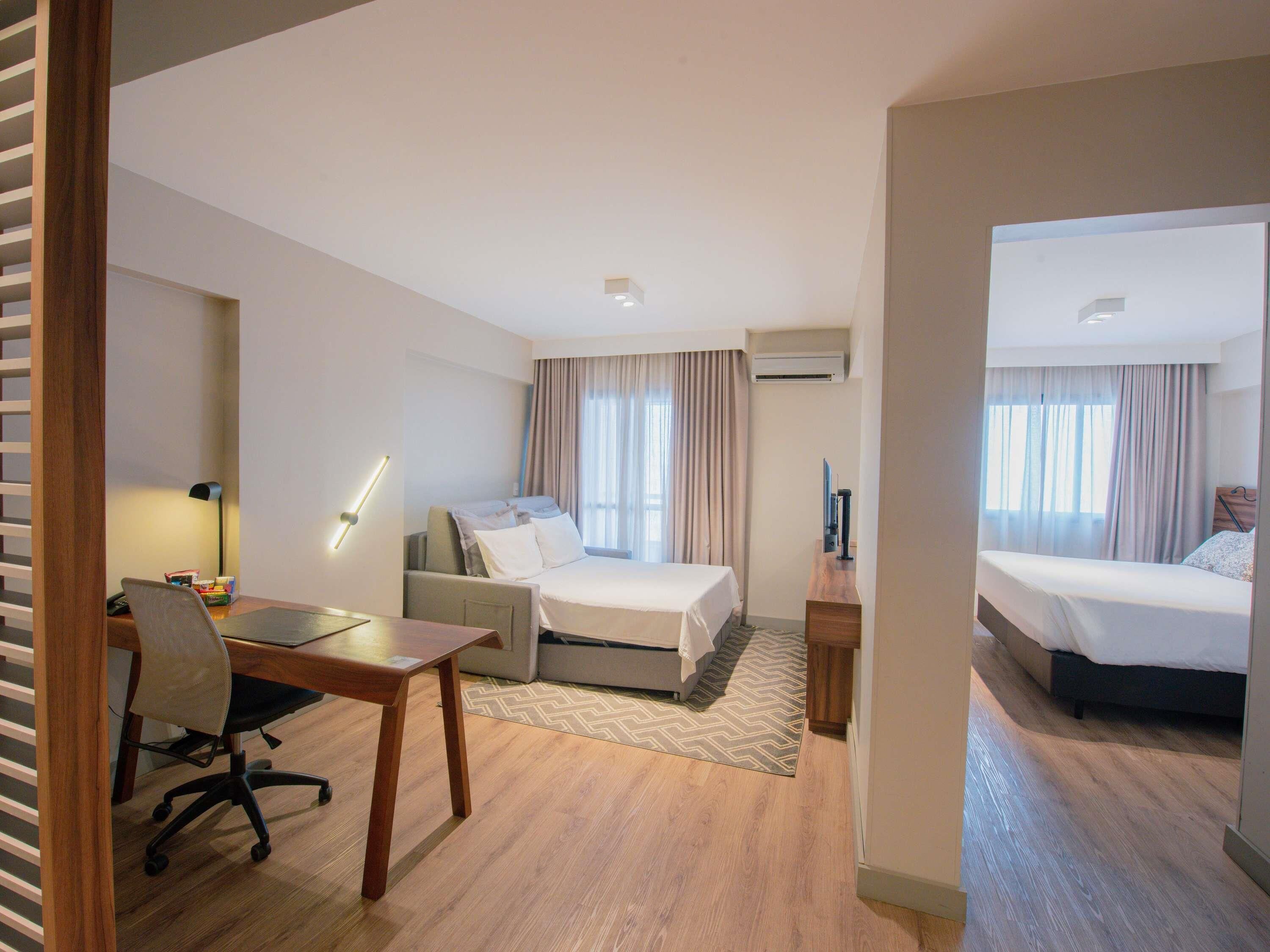 A hotel room with a desk, two beds, a chair, and a desk lamp.