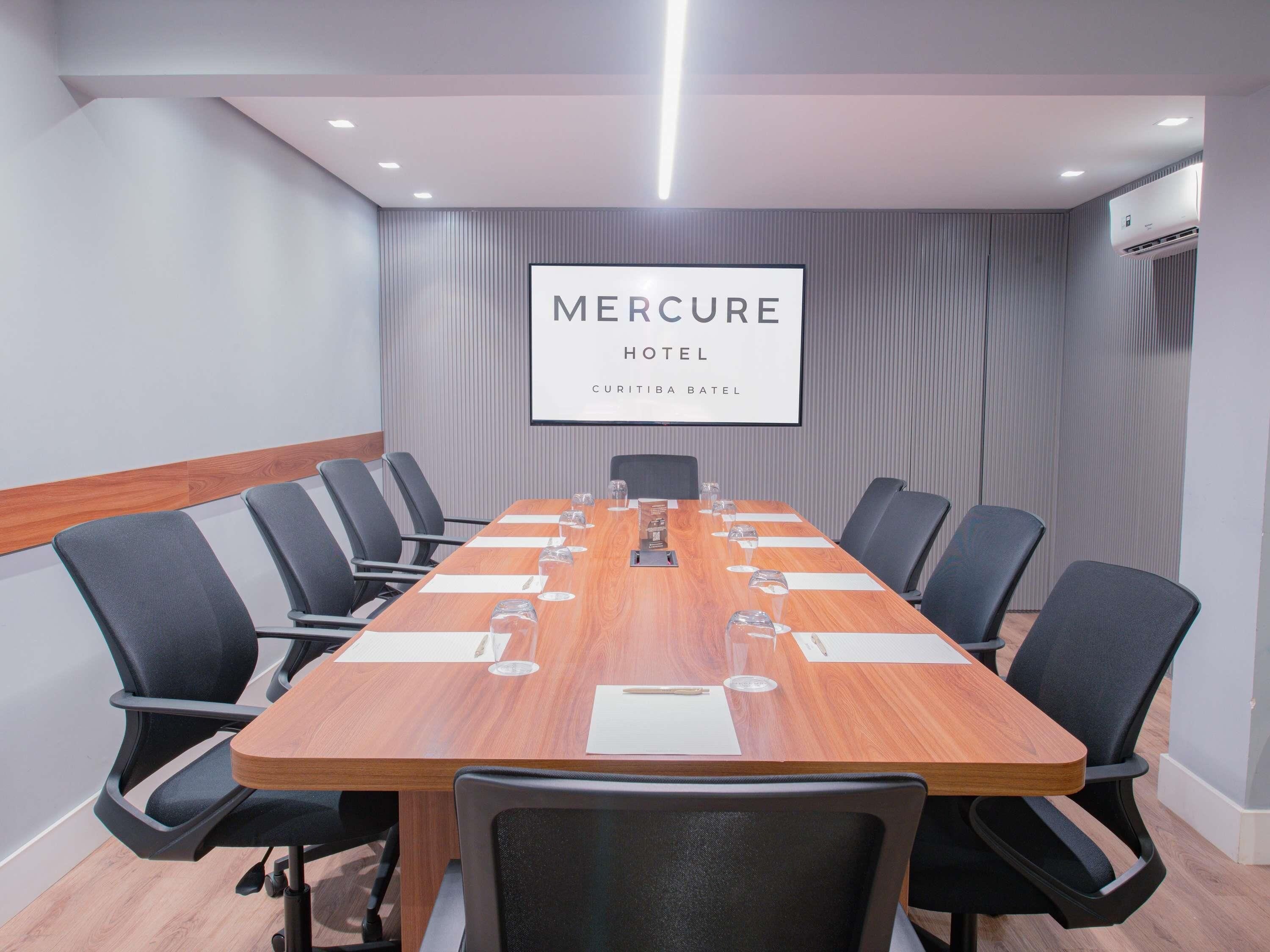 A conference room with a long wooden table, black chairs, and a large screen displaying 'Mercure Hotel Curitiba Batel'.