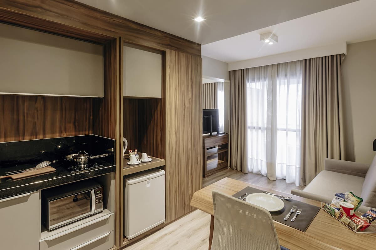 Une chambre d’hôtel moderne dotée d’une kitchenette, d’un coin repas, d’une télévision et d’une grande fenêtre avec des rideaux.