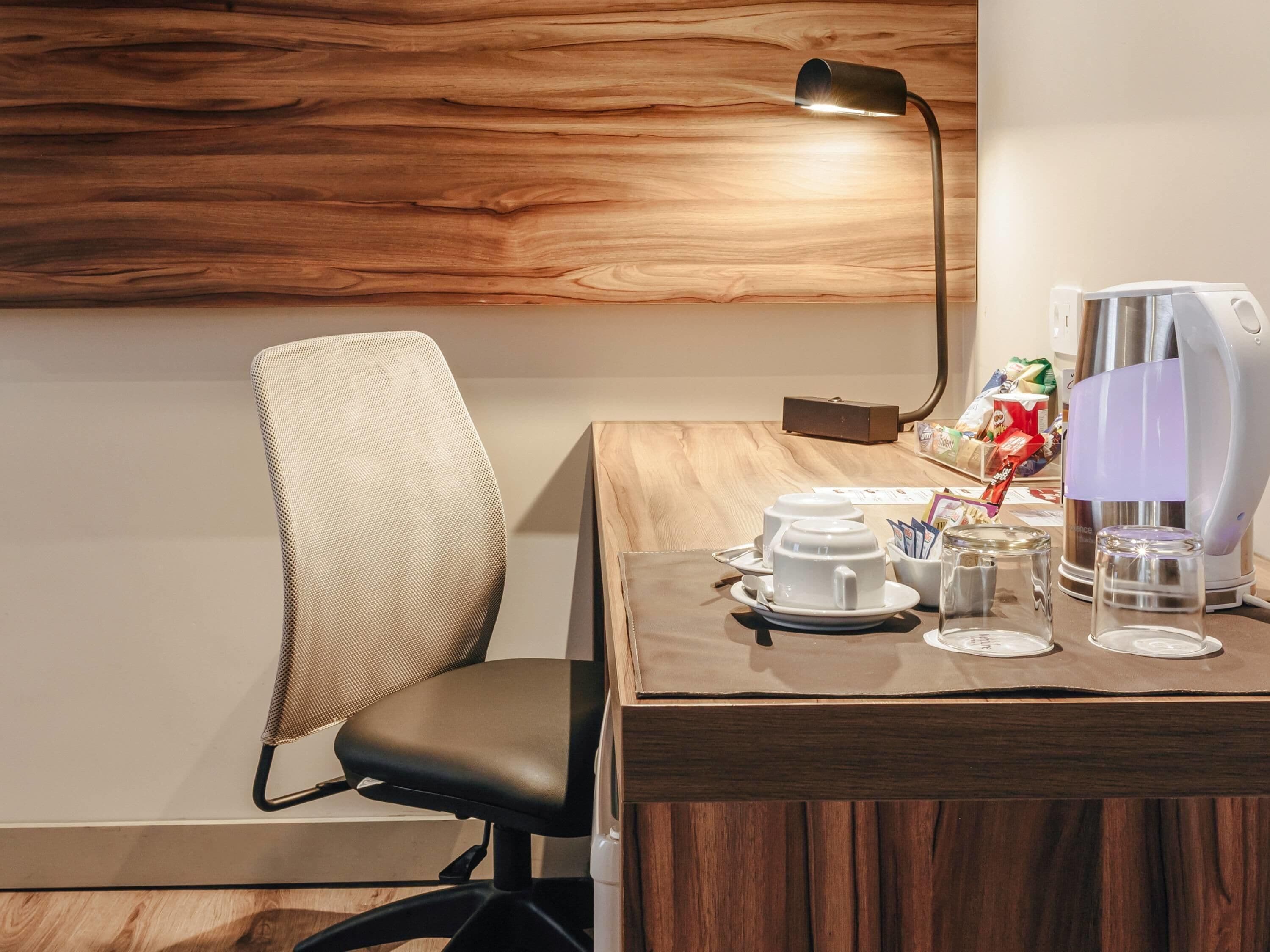 A modern hotel room with a wooden wall, a black chair, a table with a coffee machine, cups, and a kettle.