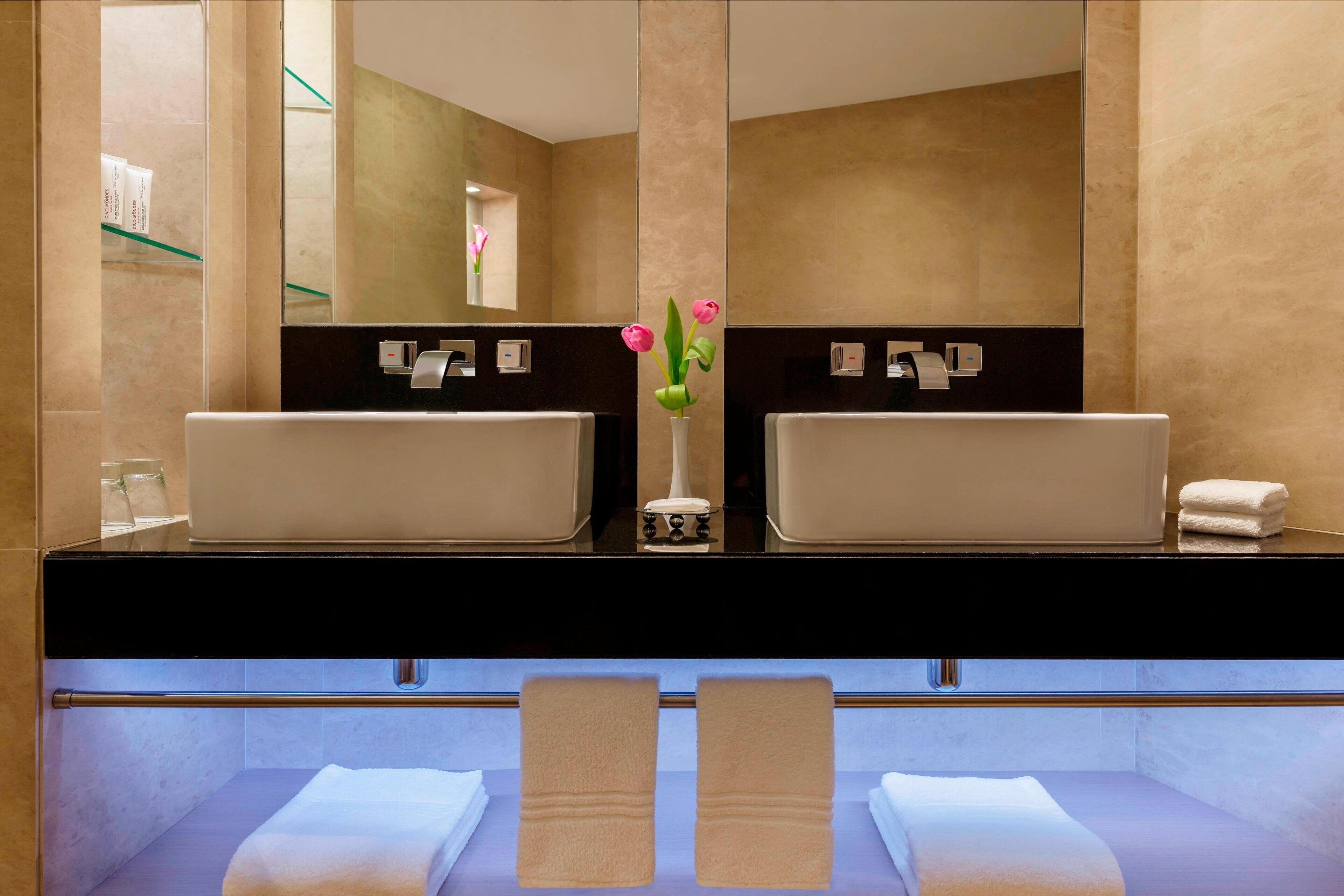 Ein Badezimmer mit zwei Waschbecken, einem Spiegel, einer Blumenvase und Handtüchern an einem Handtuchhalter.