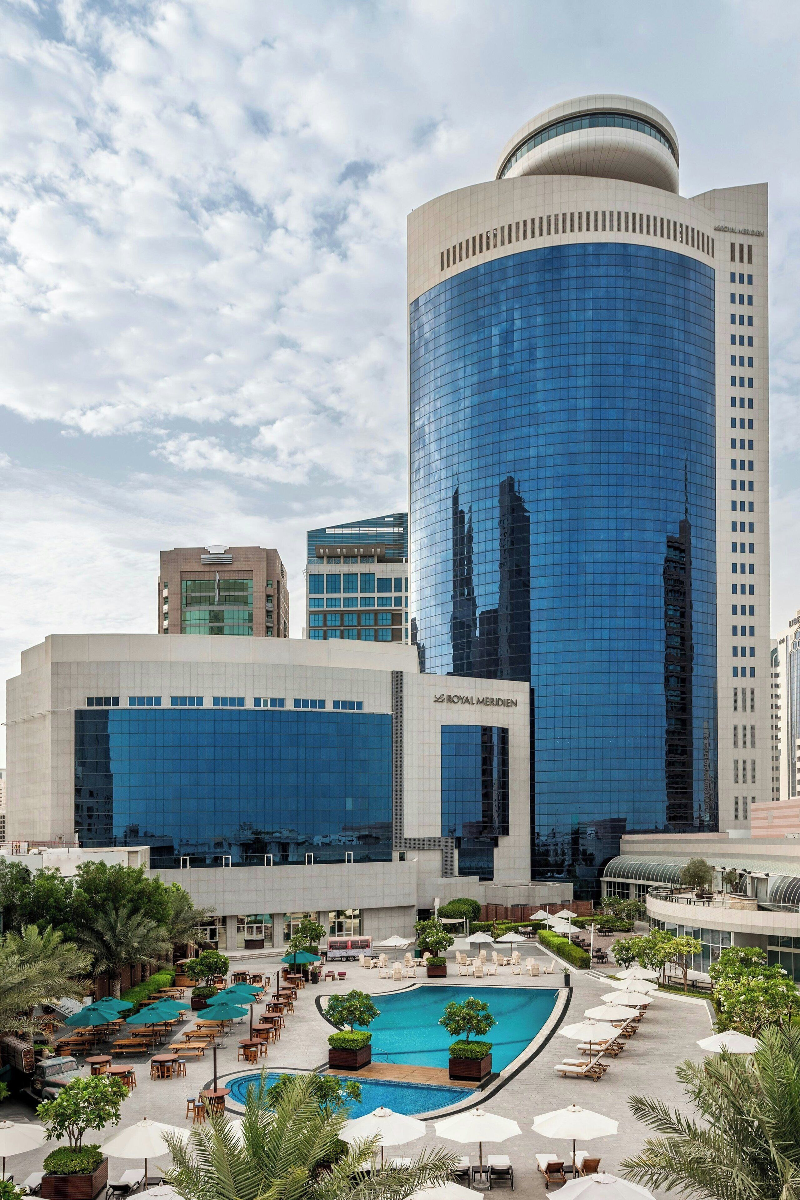 A hotel with a large pool area, outdoor seating, and a modern building with reflective glass windows.