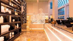 A modern lobby with marble flooring, a central seating area with black chairs, and a glass ceiling.