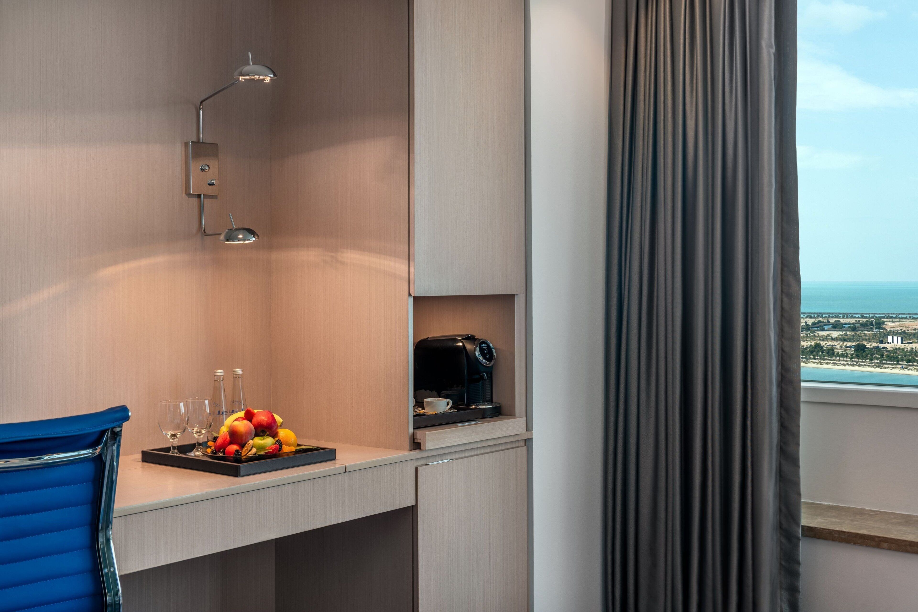 A hotel room with a desk, a coffee maker, a fruit tray, and a view of the sea.
