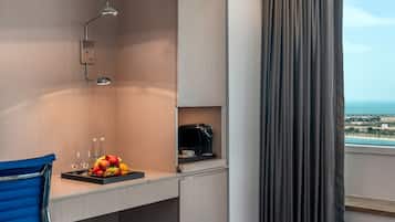 A hotel room with a desk, a coffee maker, a fruit tray, and a view of the sea.
