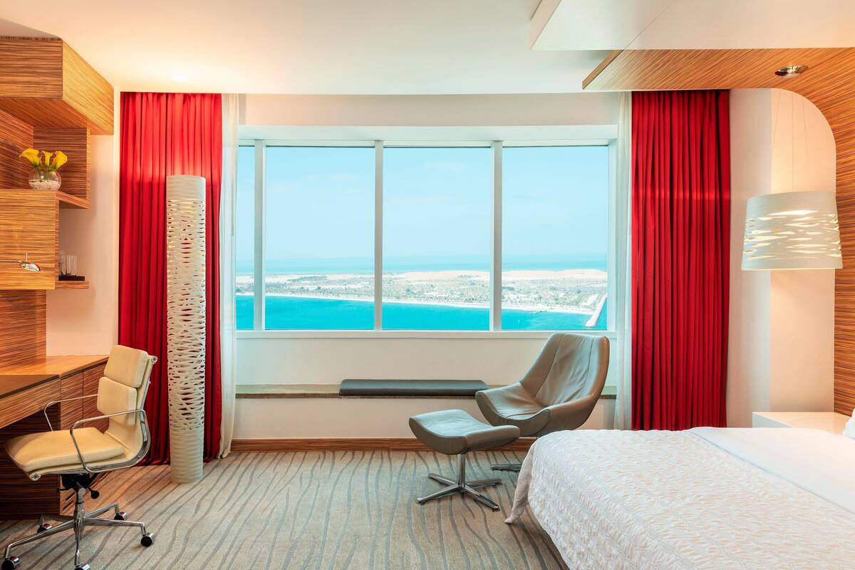 Hotelkamer met groot raam met uitzicht op strand en zee, een bed, bureau met stoel, en een rood gordijn.