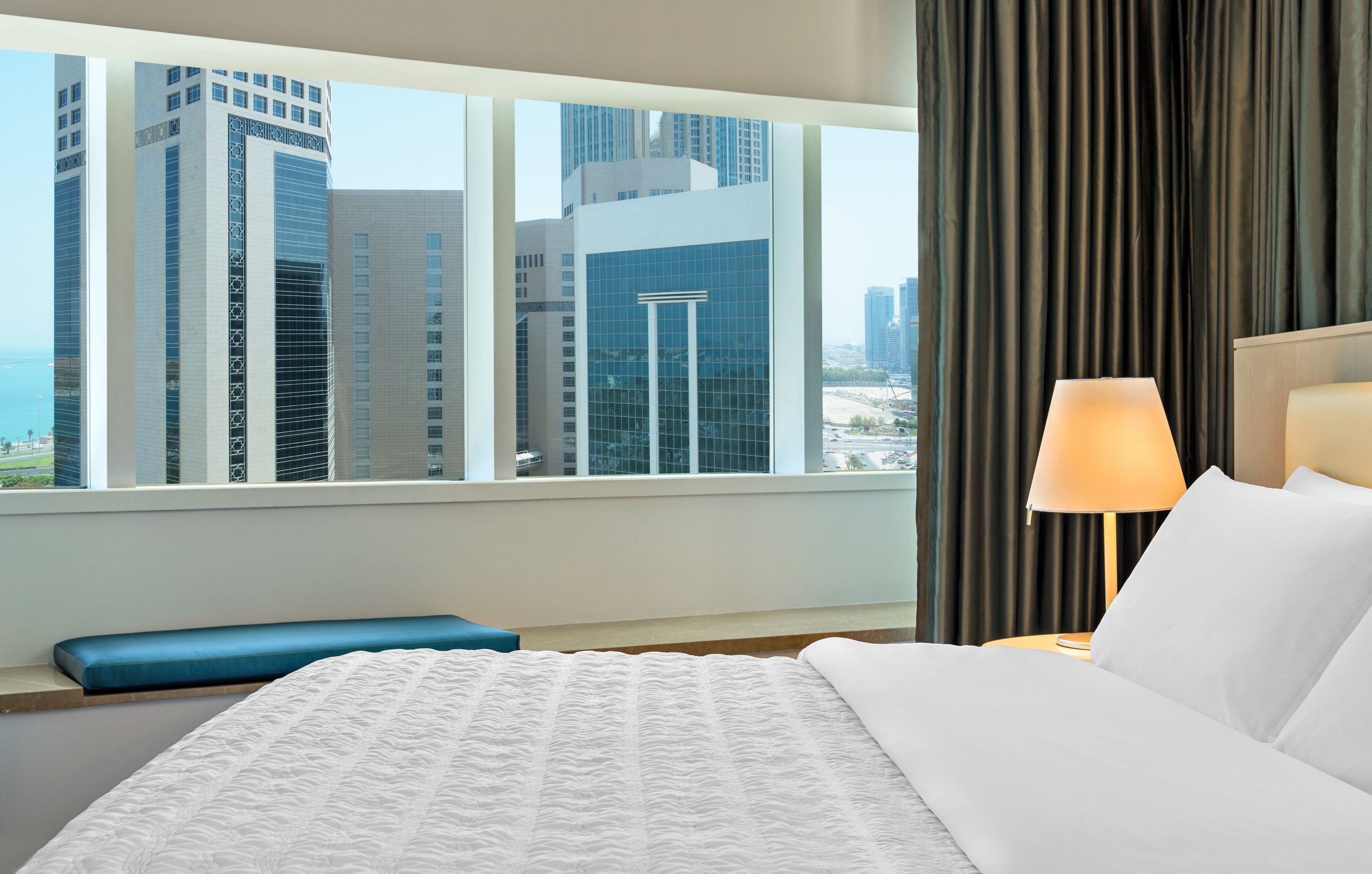 Ein Hotelzimmer mit großem Fenster und Blick auf die Stadt, einem Bett mit weißer Bettwäsche, einer blauen Bank und einer Nachttischlampe.