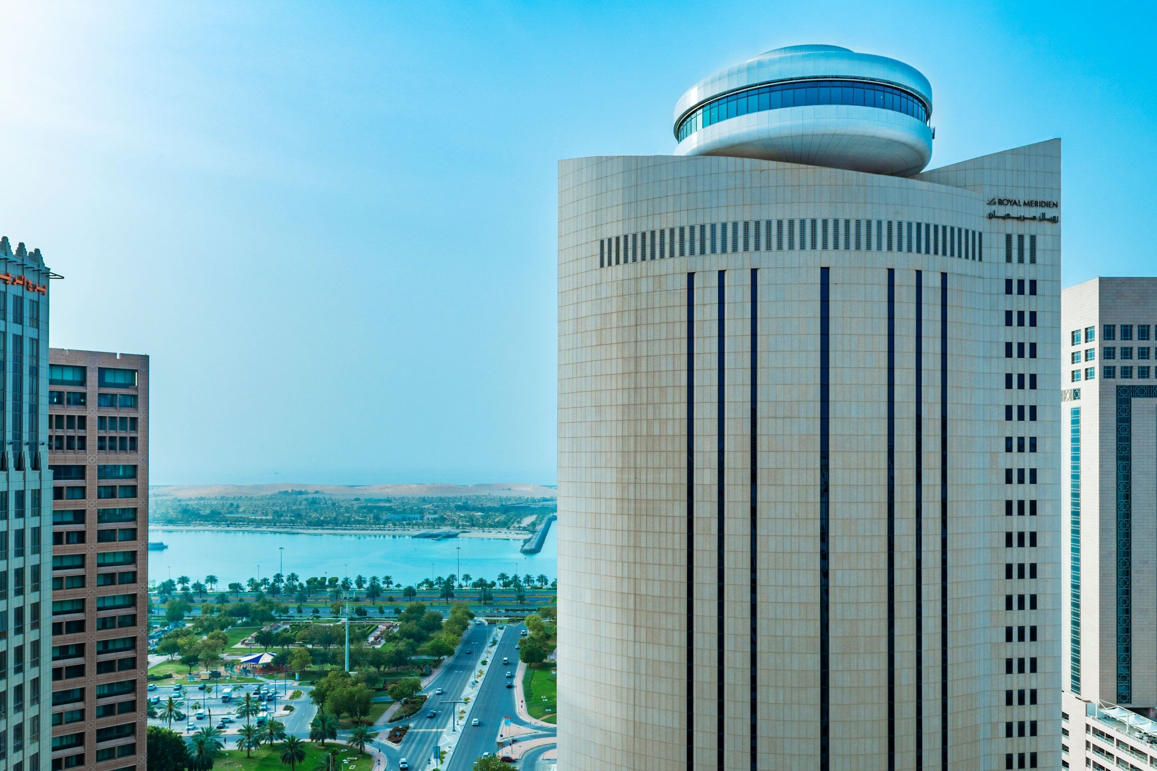 A modern high-rise building with a distinctive dome and vertical lines, surrounded by other tall buildings and a view of a coastal area.