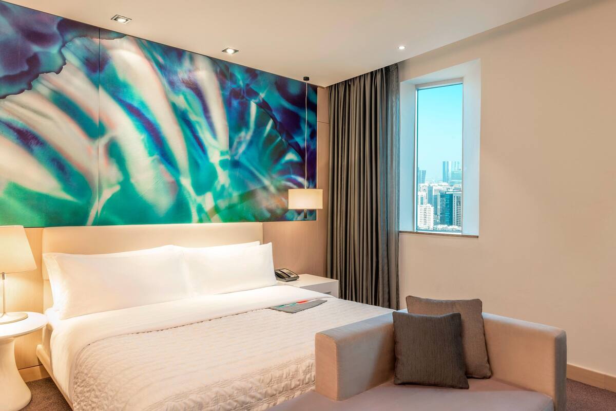 Een hotelkamer met een groot bed, een stoel, een nachtkastje, een lamp en uitzicht op de stad door een raam.