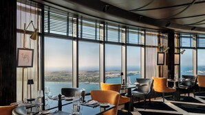 A high-rise restaurant with large windows offering a city view, modern furniture, and a black and white patterned floor.