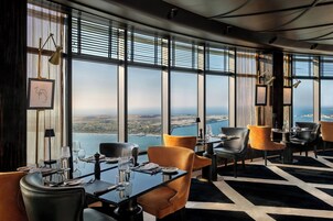 Un restaurante en un edificio alto con amplios ventanales que ofrece vistas de la ciudad, mobiliario moderno y un piso con estampado blanco y negro.