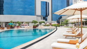 A swimming pool area with lounge chairs and umbrellas, surrounded by modern buildings with reflective glass facades.