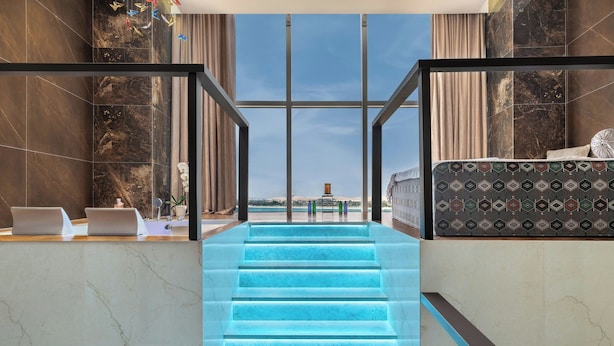 A hotel lobby with a marble reception desk, a staircase with blue lighting, and a large window offering a view of the city.
