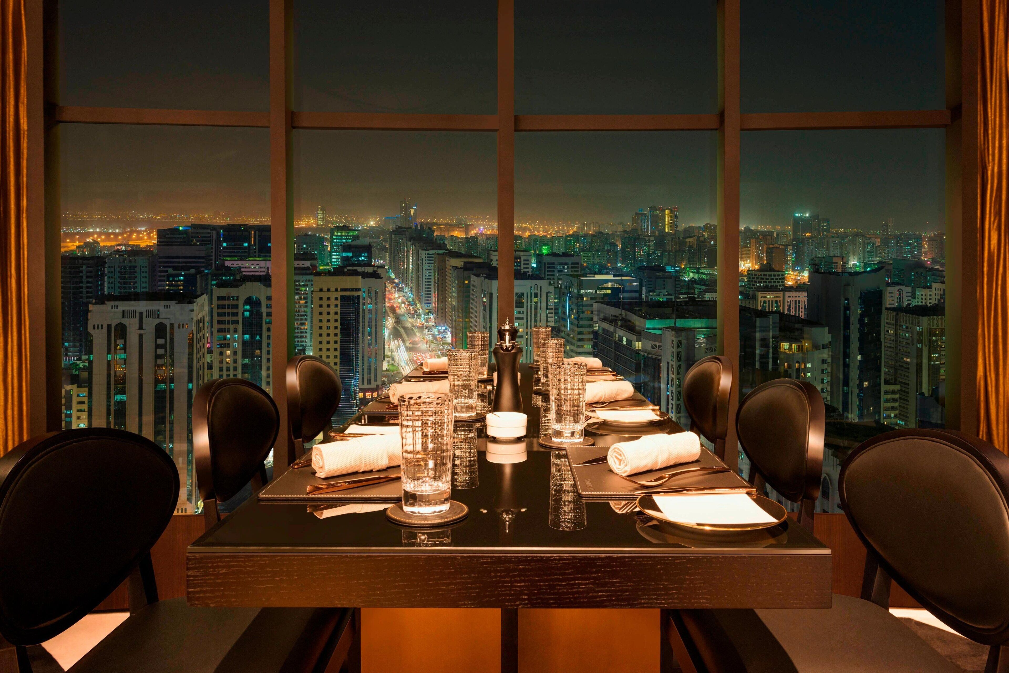 A dining table set for two with a cityscape view through large windows.