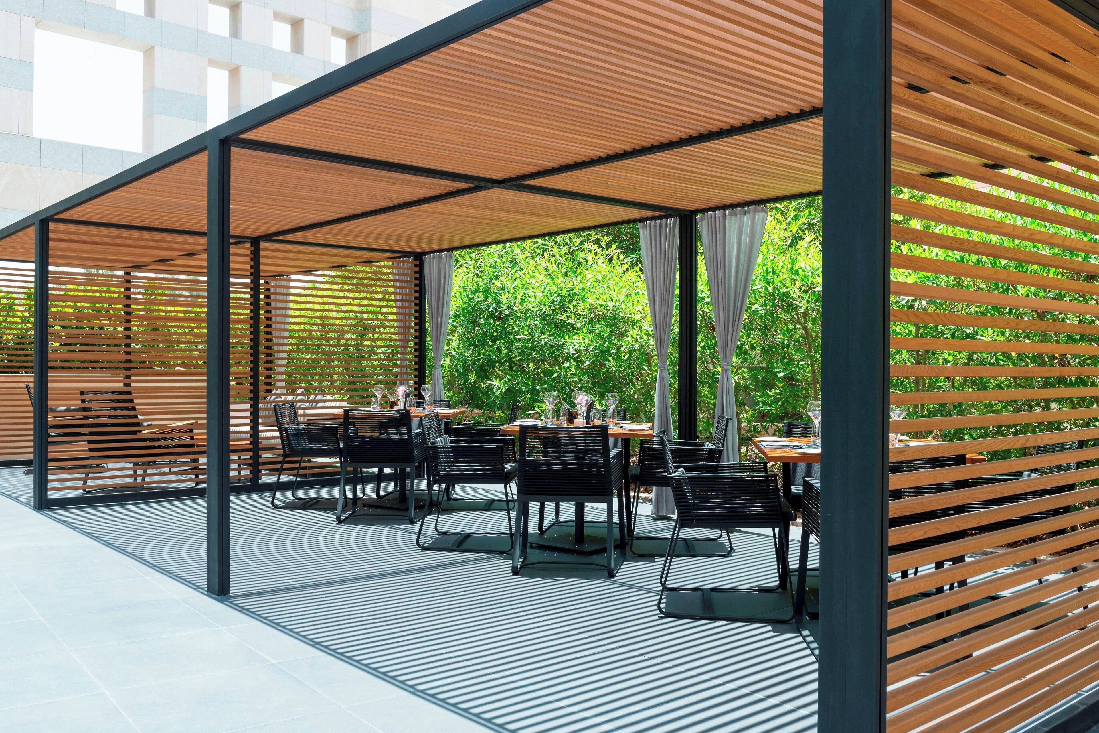 An outdoor dining area with tables and chairs, covered by a wooden pergola, and surrounded by greenery.