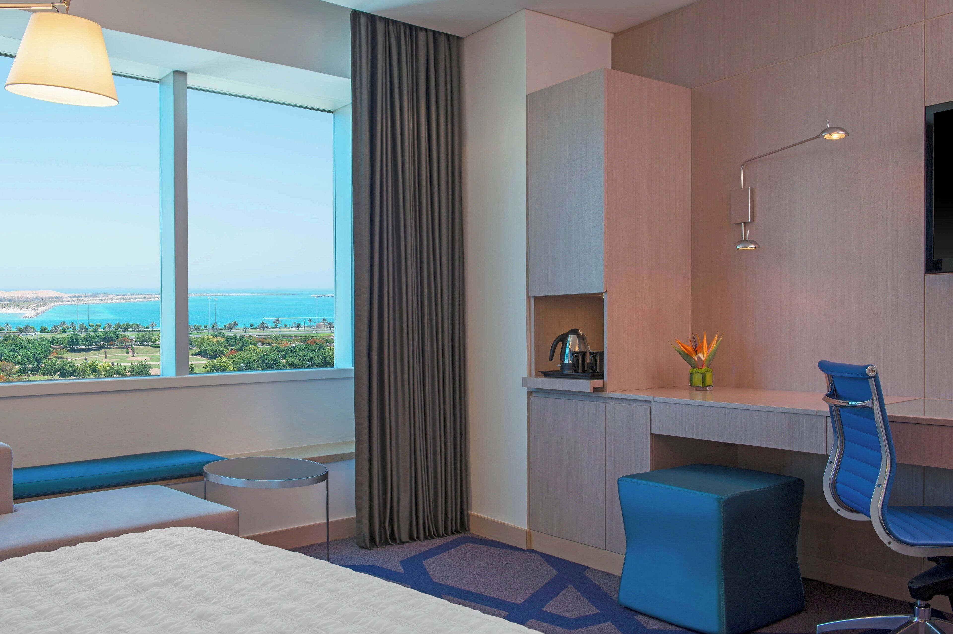 A hotel room with a large window offering a view of the sea and greenery, a desk with a chair, a small table, and a sofa.
