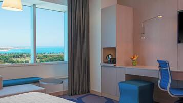 A hotel room with a large window offering a view of the sea and greenery, a desk with a chair, a small table, and a sofa.