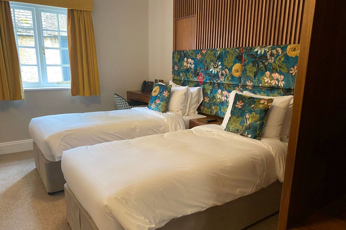 Habitación de hotel con dos camas, cabecero de madera, un escritorio pequeño y una ventana con cortinas.