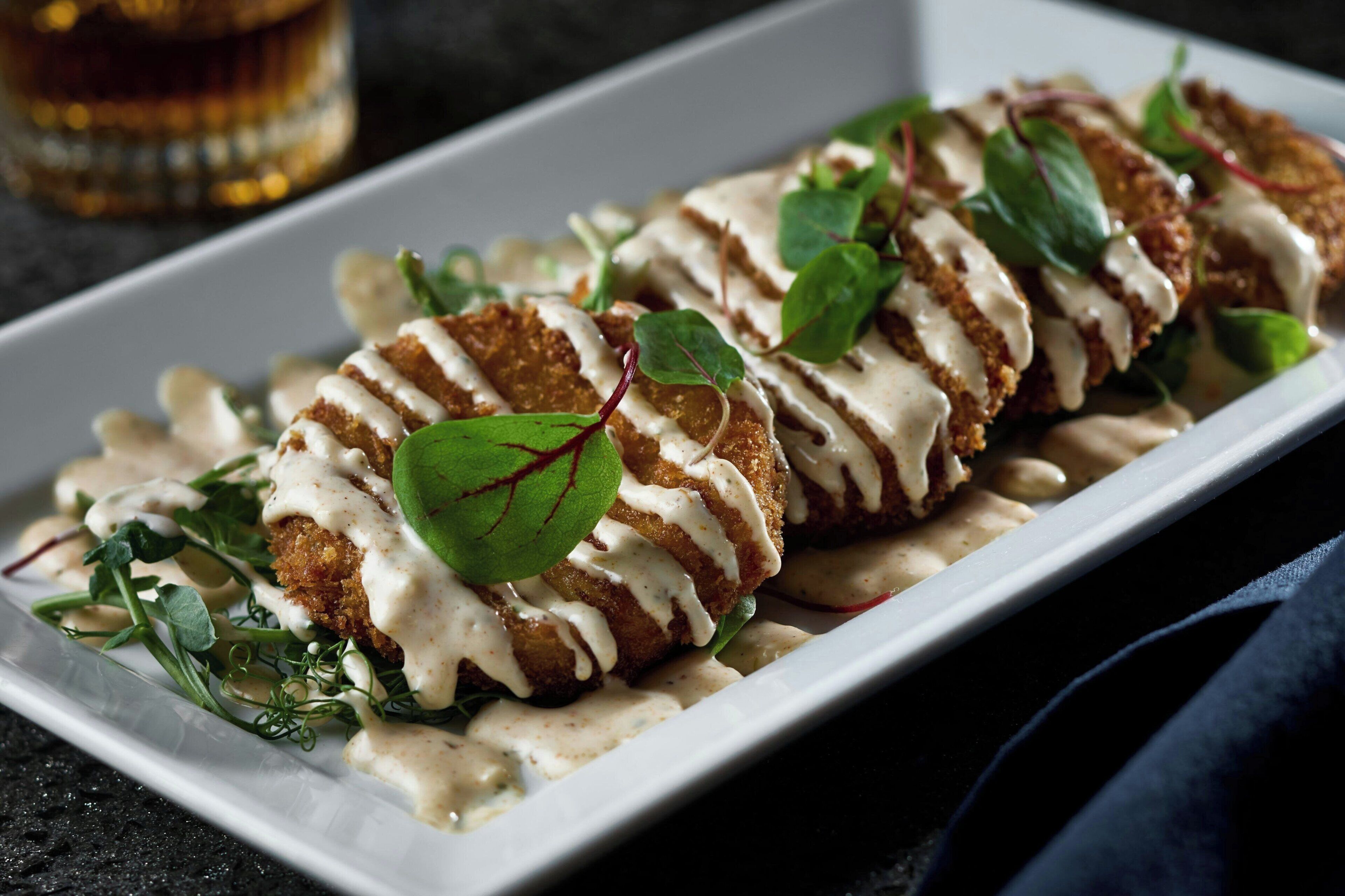 Une assiette rectangulaire blanche sur laquelle est présentée une préparation culinaire nappée d’une sauce crémeuse, agrémentée de feuilles vertes, le tout sur un fond sombre.