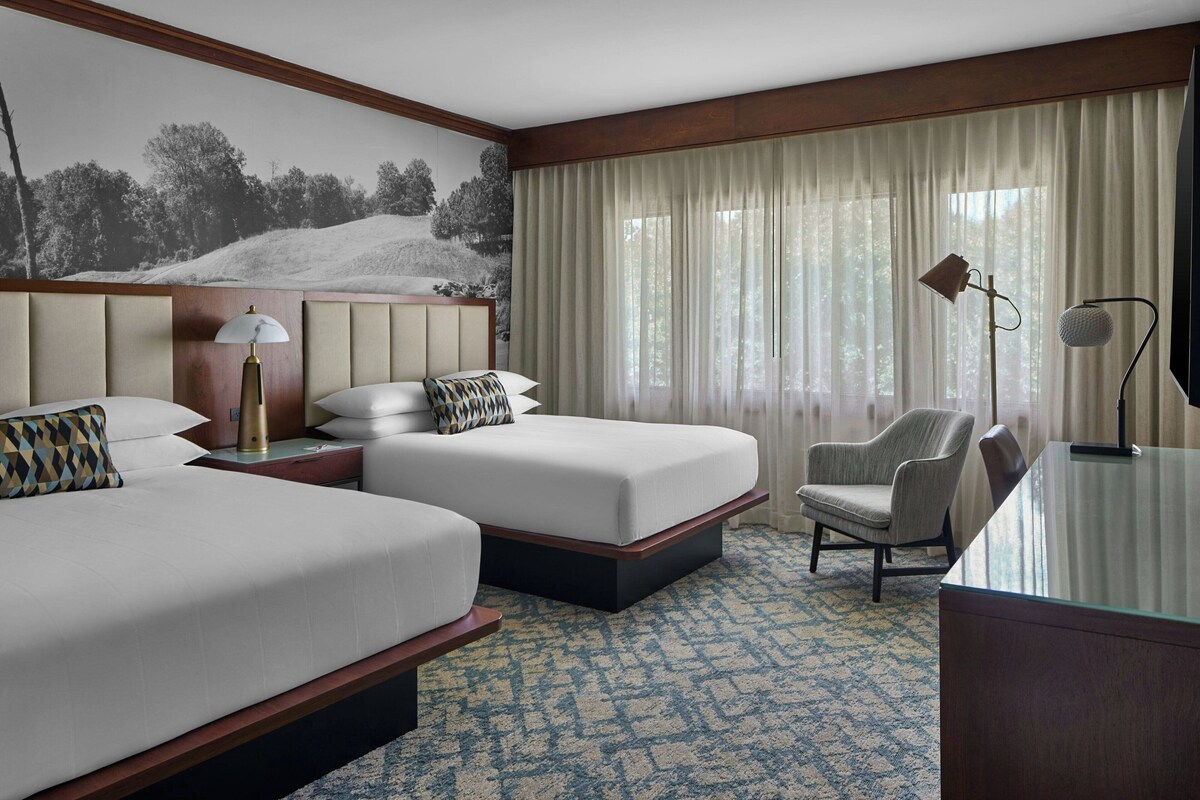 A hotel room with two beds, a desk, a chair, and a large window with curtains.