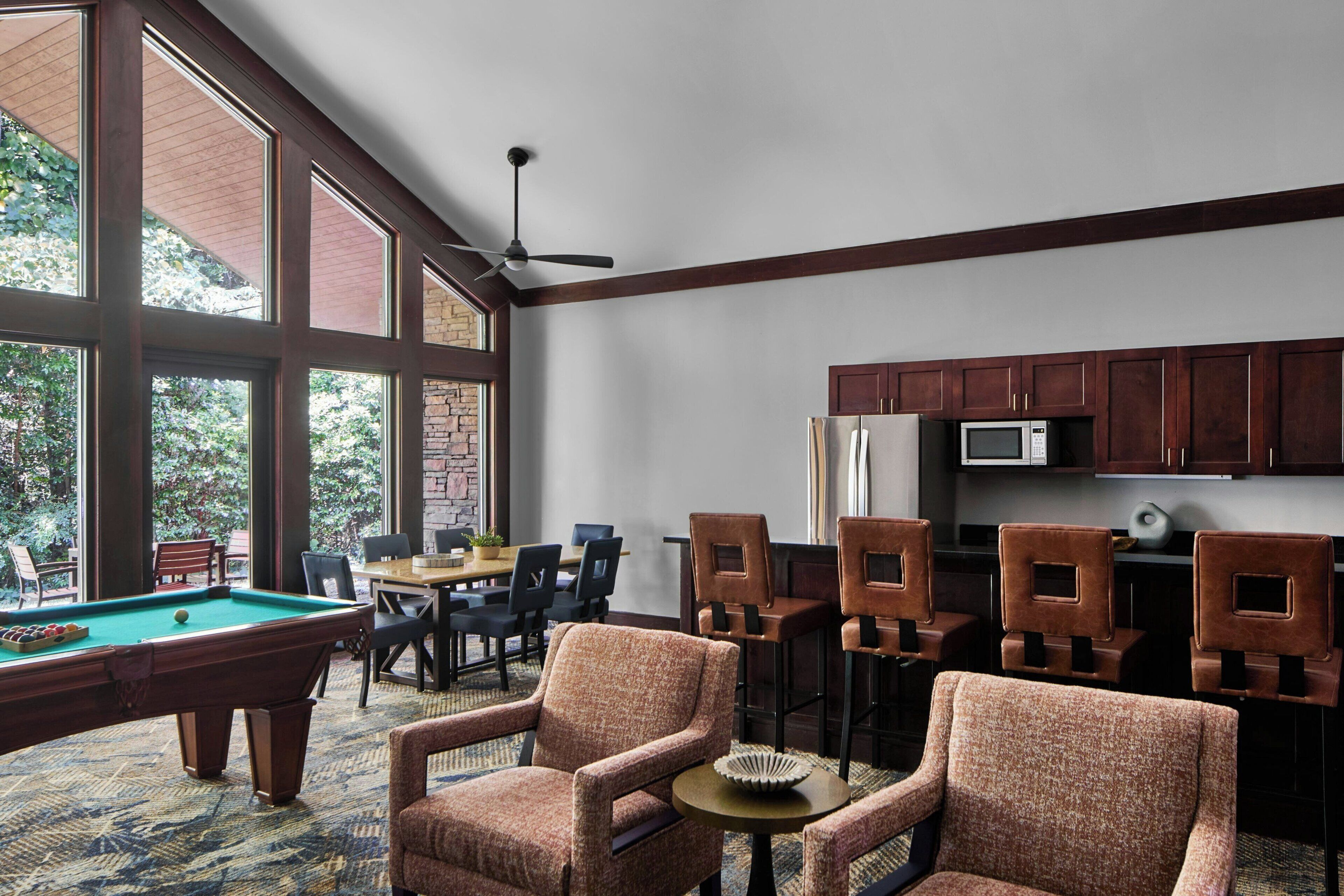 Una mesa de billar en una habitación con ventanas amplias, un ventilador de techo y una zona de cocina con armarios de madera.