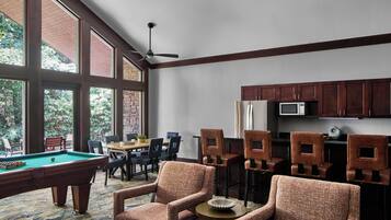 Una mesa de billar en una habitación con ventanas amplias, un ventilador de techo y una zona de cocina con armarios de madera.