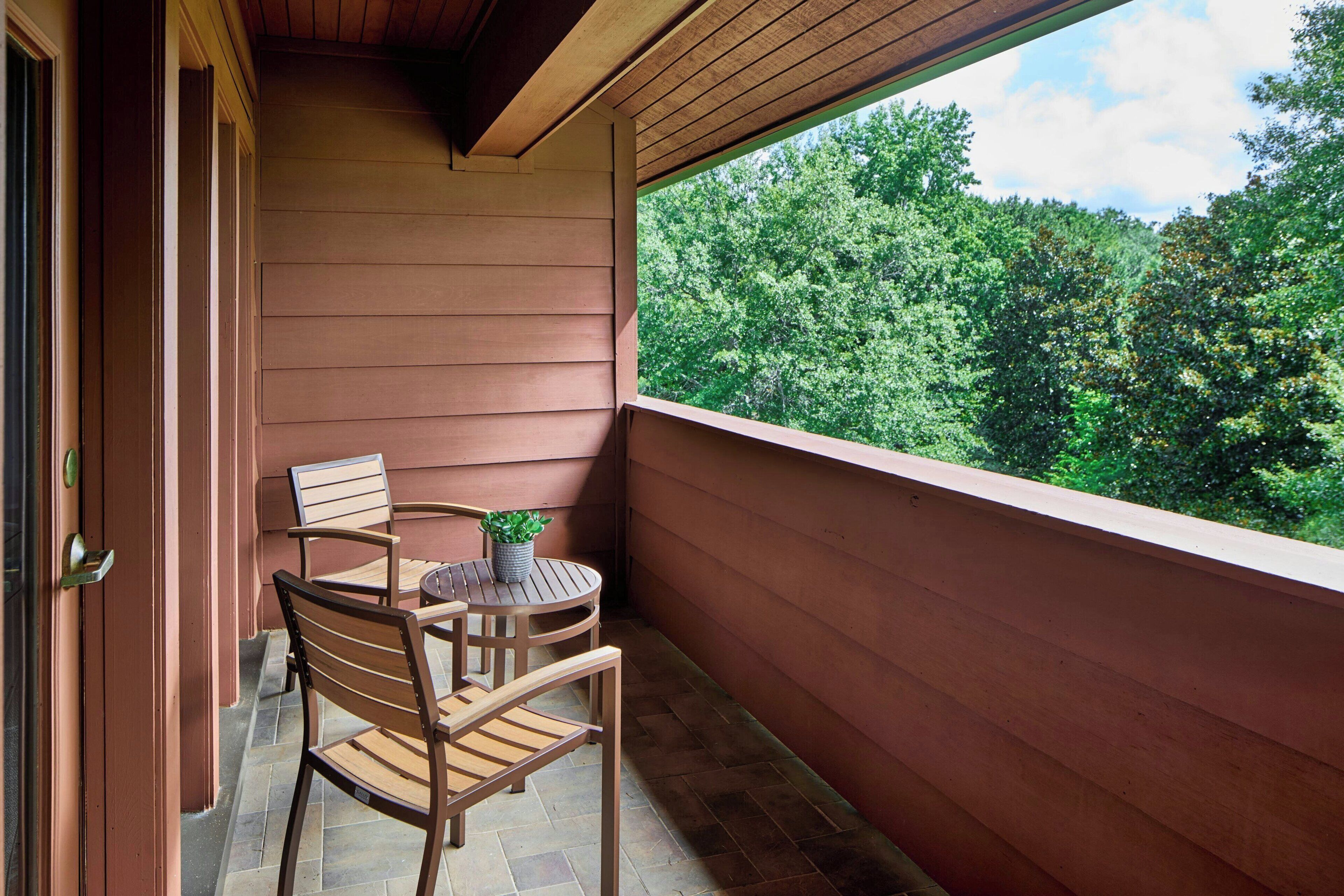 A balcony with wooden chairs and a table, a potted plant, and a view of a forest.