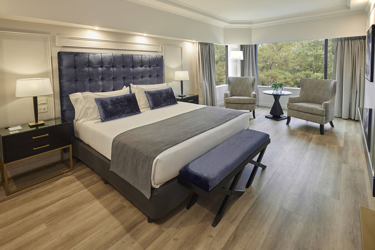 Een moderne hotelkamer met een groot bed, nachtkastjes, een bankje, twee fauteuils, een tafeltje met een plant en uitzicht op bomen door het raam.