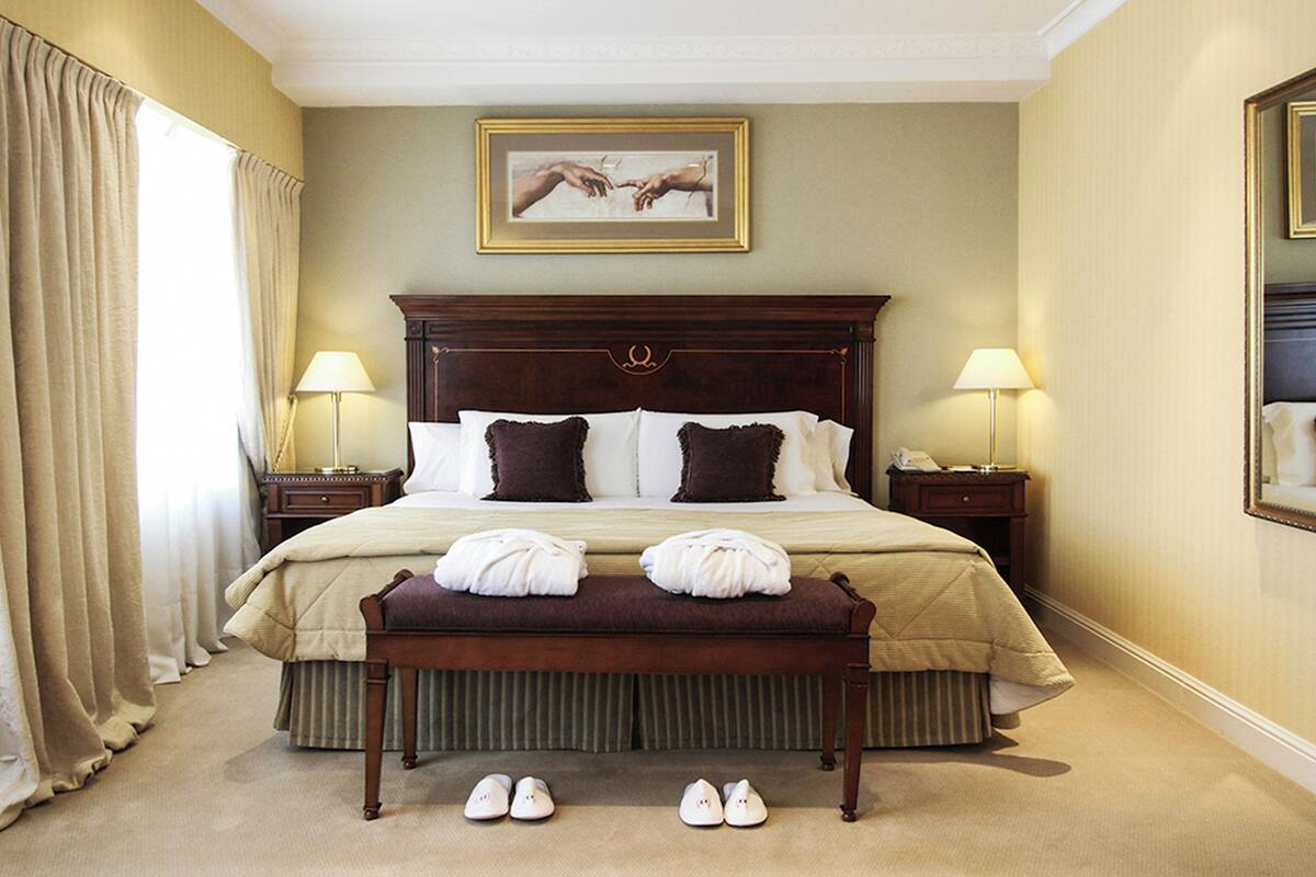 Een hotelkamer met een groot bed, twee nachtkastjes, een bankje en een paar schoenen op de vloer.