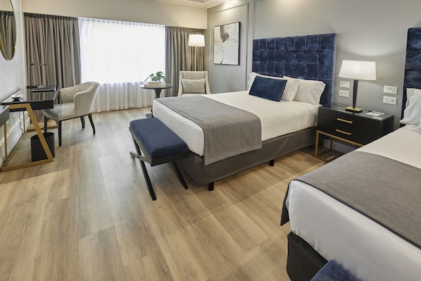 A hotel room with a large bed, a desk, a chair, and a small table with a lamp.