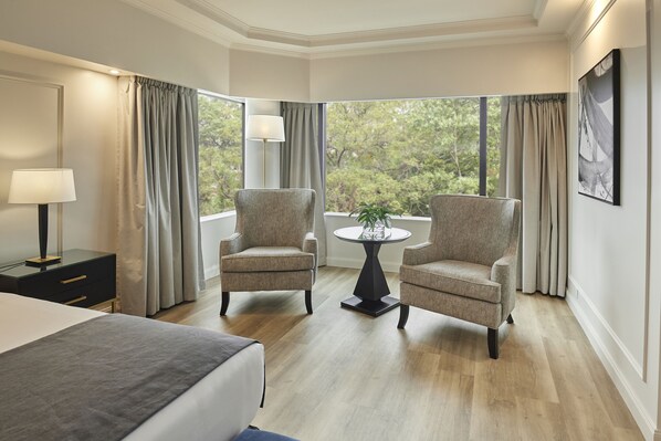 A hotel room with a bed, two armchairs, a small table with a plant, a lamp, and a large window with curtains.