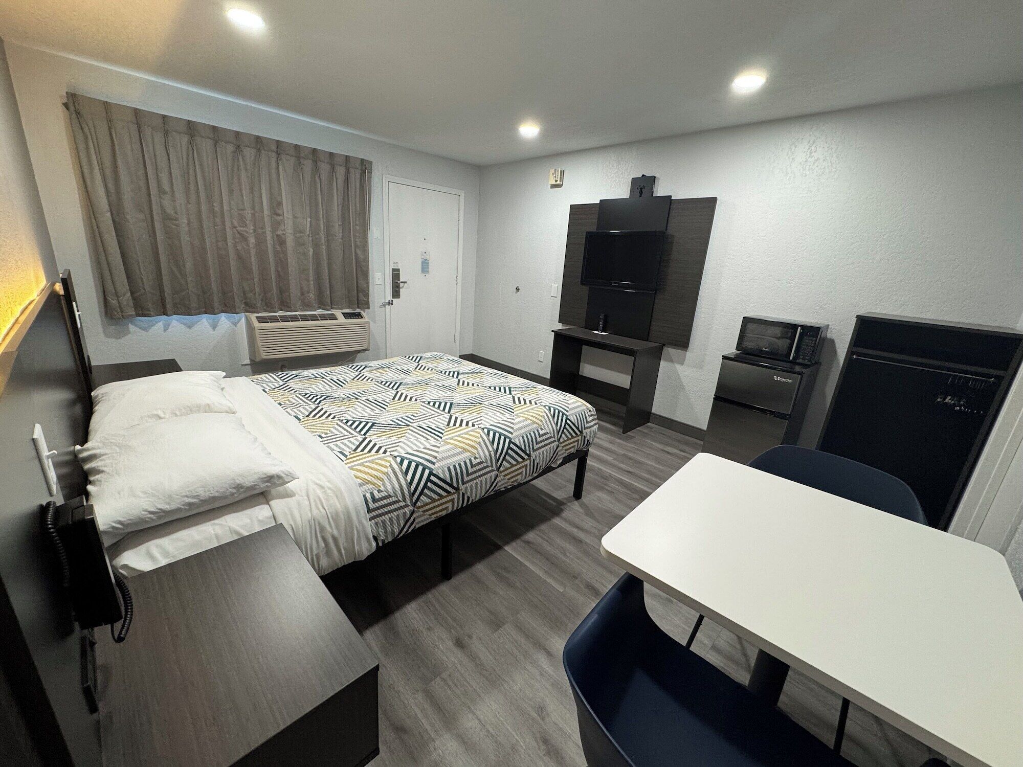 A modern hotel room with a bed, bedside table, desk, chair, TV, and a minibar.