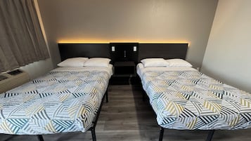 A hotel room with two single beds, each with a patterned bedspread and white pillows. There is a nightstand with a lamp between the beds, and the room has a neutral color scheme with a wooden floor.