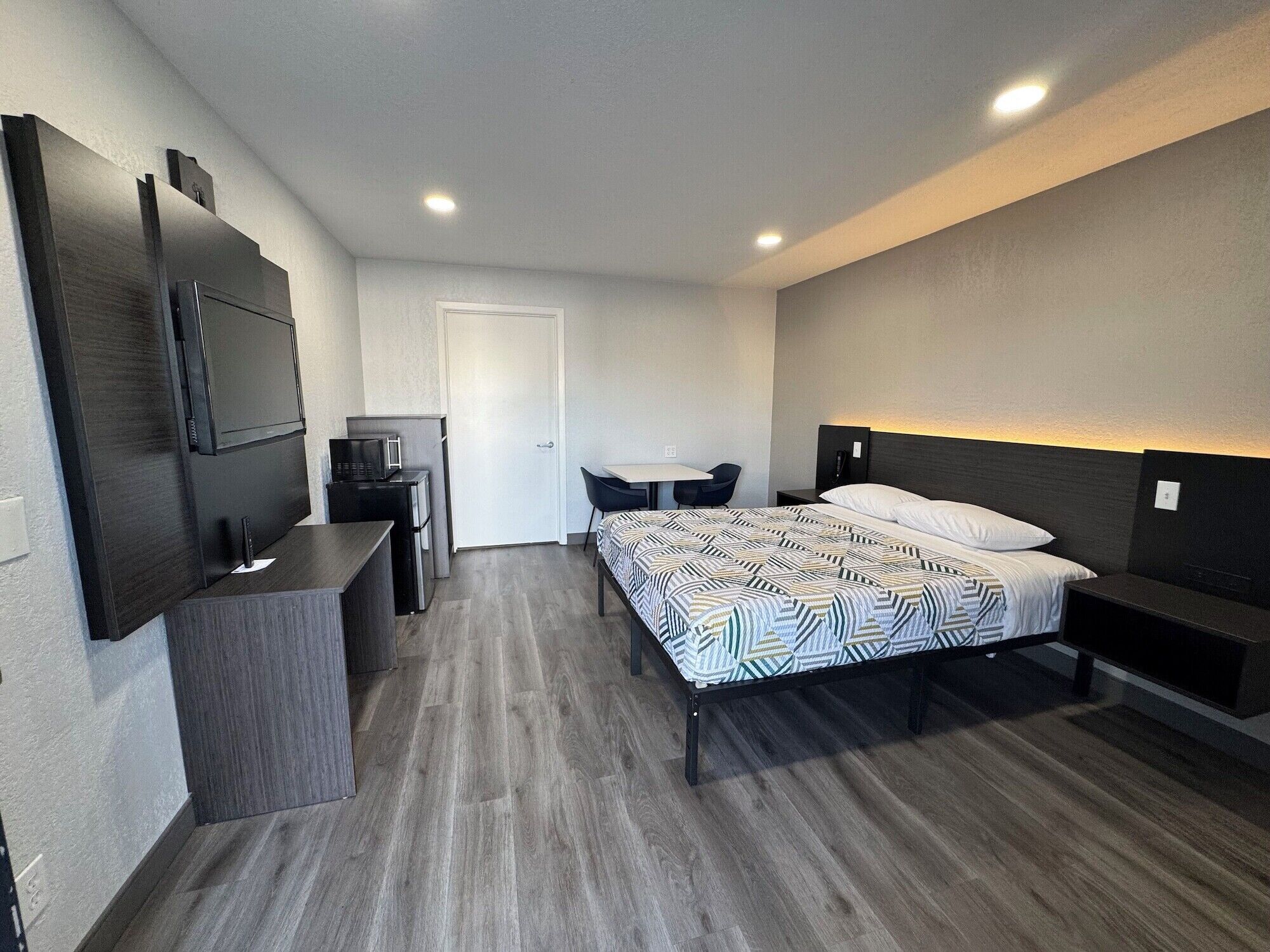 Una habitación de hotel compacta con cama, mesitas de noche, escritorio con silla, televisor y refrigerador.