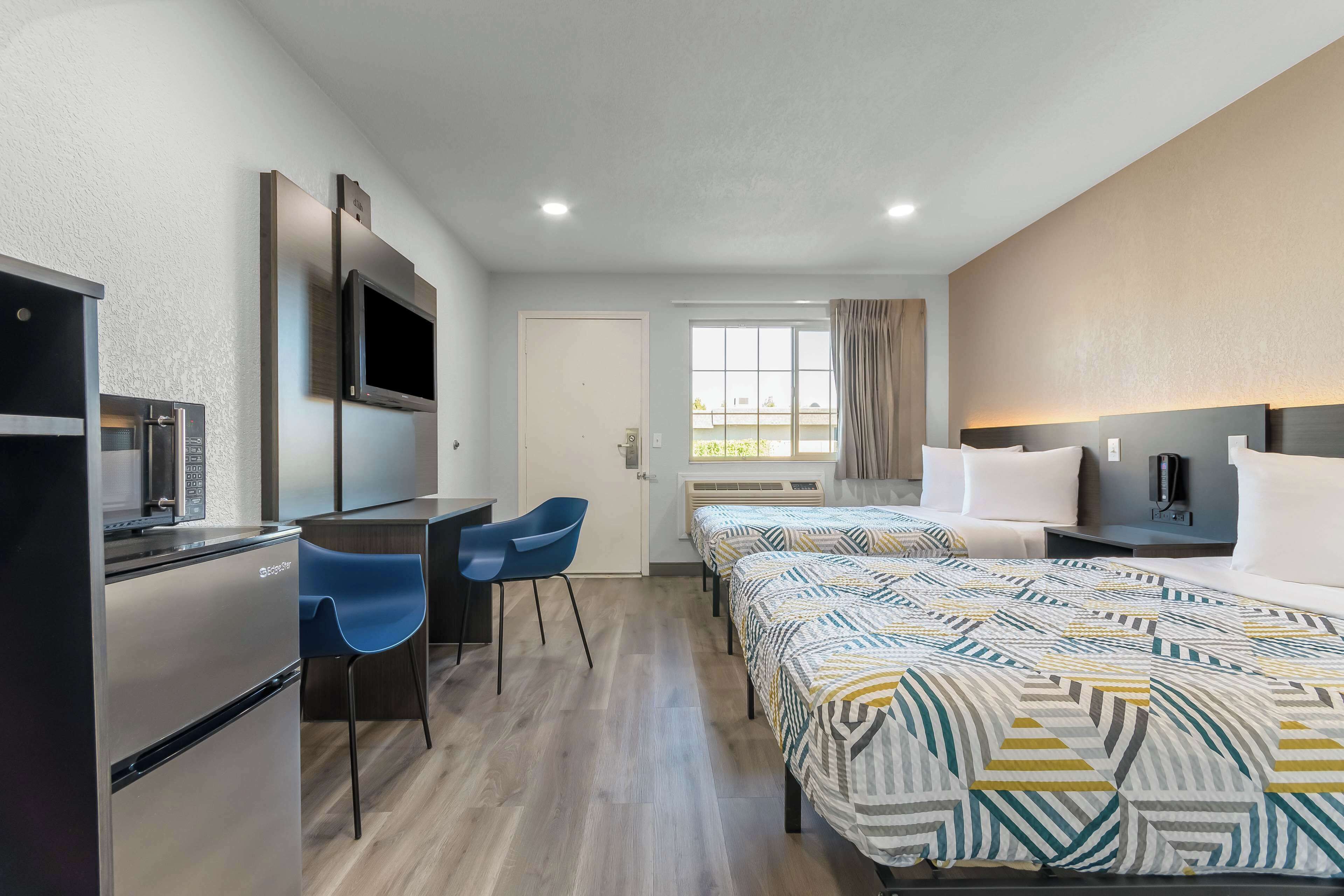 A hotel room with a kitchenette, two beds, a TV, and a window with curtains.