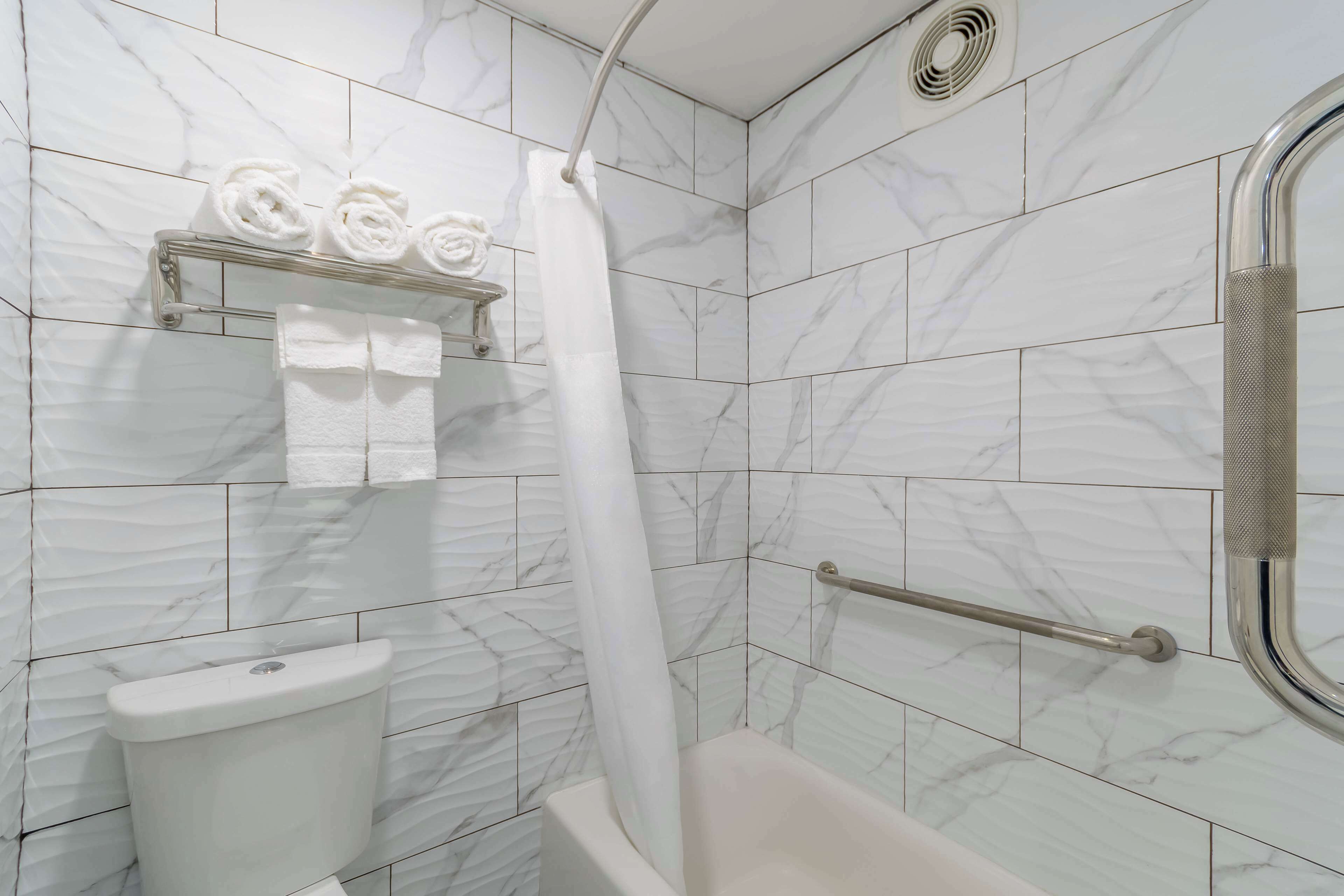 A bathroom with a white toilet, a bathtub, and a shower with a curtain. There is a towel rack with towels and a handrail for the shower.