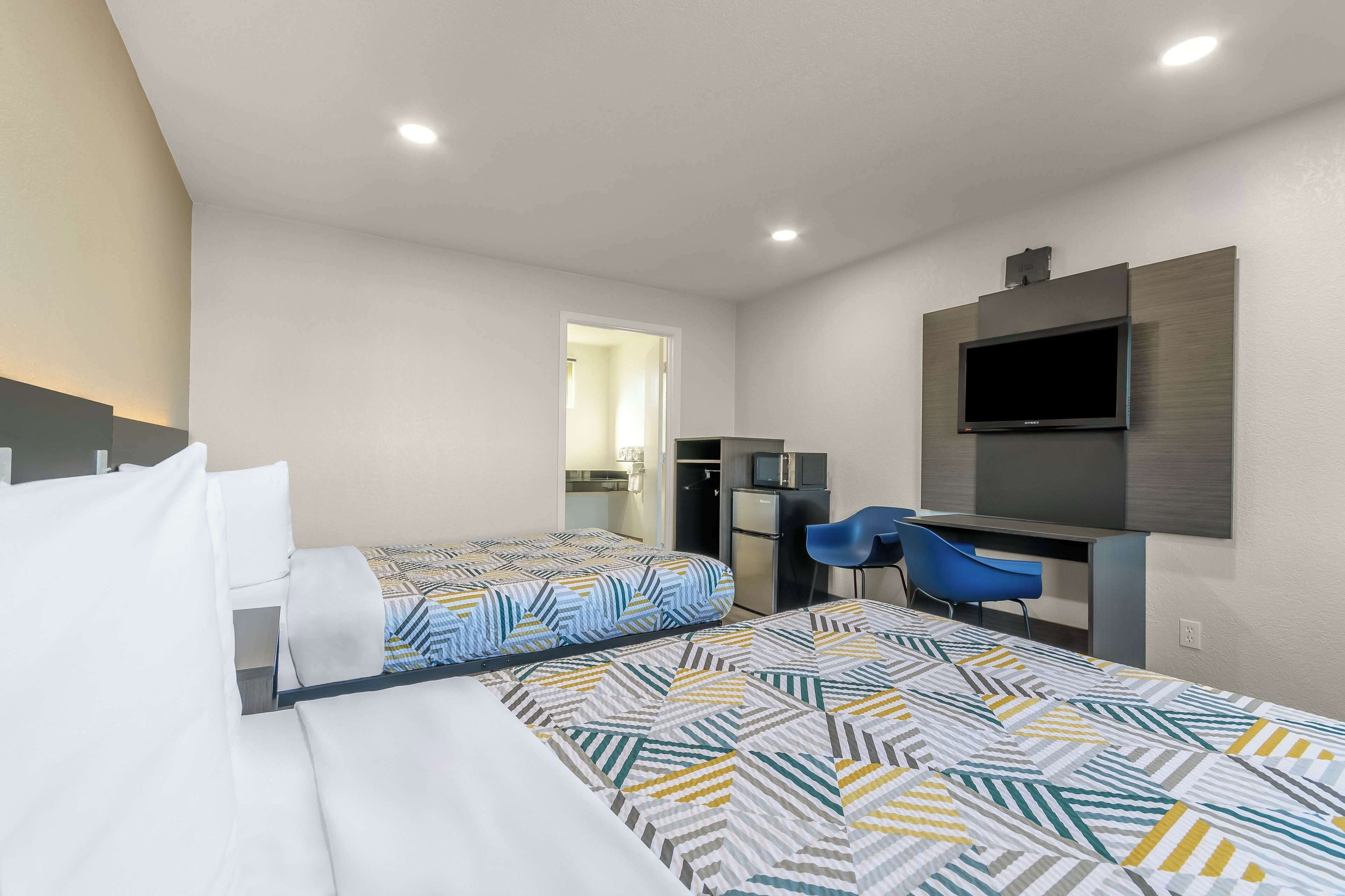 A hotel room with two beds, a desk, a chair, a TV, and a kitchenette.